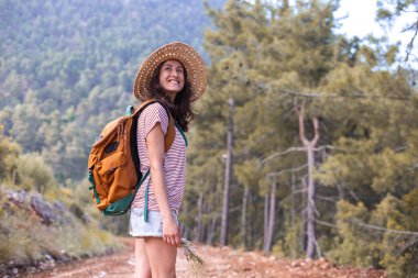 Ormanda sırt çantası ve hasır şapkalı bir kadının portresi, yürüyüş yapan gülümseyen bir kız, yaz yürüyüşü yapan, dağ yolunda yürümekten zevk alan mutlu bir kadın.
