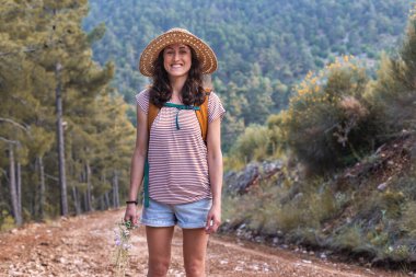 Ormanda sırt çantası ve hasır şapkalı bir kadının portresi, yürüyüş yapan gülümseyen bir kız, yaz yürüyüşü yapan, dağ yolunda yürümekten zevk alan mutlu bir kadın.