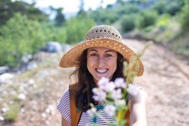 Hasır şapkalı bir kız elinde bir buket yaban çiçeği tutuyor, gülümseyen bir kadın elinde buket tutuyor, doğada bir yürüyüş.