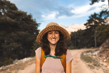 Dağlarda yürüyüşe çıkmış sırt çantalı bir kadın, orman yolunda yürüyen şapkalı bir kız, gülümseyen bir esmer