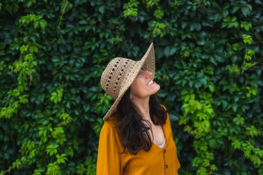 Hasır şapkalı gülümseyen bir kız sarmaşıkla kaplanmış bir duvarın yanında duruyor. Siyah saçlı bir kadının portresi.