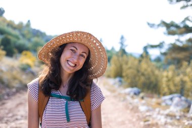 Ormanda sırt çantası ve hasır şapkalı bir kadının portresi, yürüyüş yapan gülümseyen bir kız, yaz yürüyüşü yapan, ormanda gezmekten zevk alan mutlu bir kadın.
