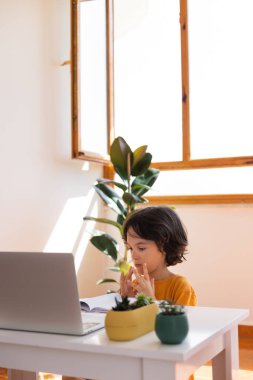 Uzaklık Eğitimi, okul çocuğunun portresi, öğretmenle video görüşmesi, çocuk masaya oturur ve parmaklar, jestler için ısınır, çocuk dizüstü bilgisayara bakar.