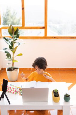 Uzaklık Eğitimi, okul çocuğunun portresi, öğretmenle video görüşmesi, çocuk masaya oturur ve parmaklar, jestler için ısınır, çocuk dizüstü bilgisayara bakar.