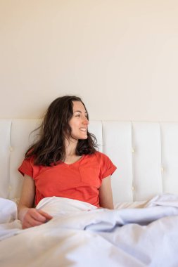 A girl in red pajamas sits on a white bed, morning awakening, portrait of a smiling woman