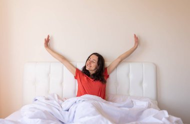 Kız yatağa oturur ve gerilir, kadın uyanır, beyaz bir yatakta oturan kırmızı tişörtlü esmer bir kadın portresi