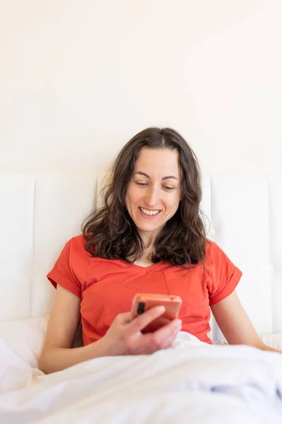 A woman sits on a bed and holds a smartphone in her hands, the girl woke up and lies on the bed with a phone