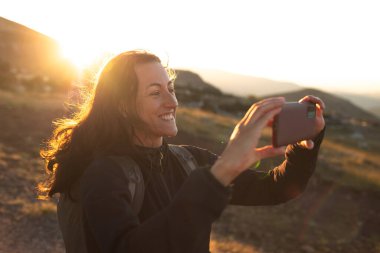 Güzel genç kız seyahat eder ve telefonda fotoğraf çeker. Dağlarda dolaşmak. Gün batımında elinde telefon olan bir kızın portresi..