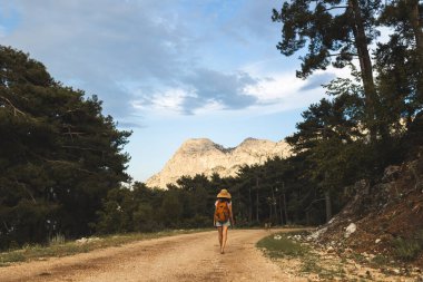 Sırt çantası ve hasır şapkalı bir kız büyük bir dağın arka planında, genç bir kadın dağ yolunda yürüyor, dünyanın en güzel yerlerine doğru bir yolculuk.