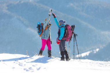 two women climbed to the top of the mountain during a winter hike, winter trekking, two girlfriends travel together, snow-capped mountains, girl gives high five to friend