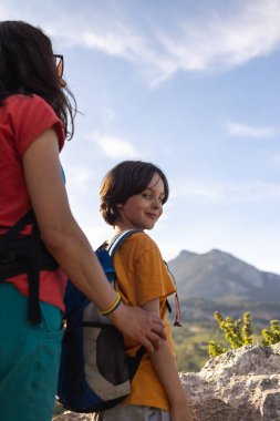 Bir kadın ve bir çocuk şafak vakti bir dağın tepesinde, dağların üzerinden yükselen güzel bir güneş manzarası, çocuklarla seyahat ediyor, anne oğluyla aktif olarak vakit geçiriyor, çocuk annesine sarılıyor.