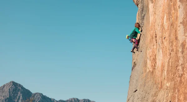 A woman climbs a rock against a blue sky, a strong girl trains strength and endurance, an extreme sport, rock climbing on natural terrain, a rock climber climbs with a rope