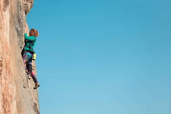 A woman climbs a rock against a blue sky, a strong girl trains strength and endurance, an extreme sport, rock climbing on natural terrain, a rock climber climbs with a rope