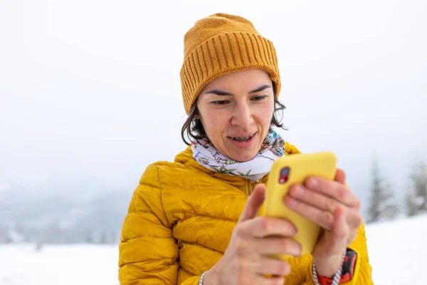 a woman in a yellow jacket holds a yellow smartphone in her hands, a girl takes a selfie against the backdrop of snow-capped mountains, winter trekking