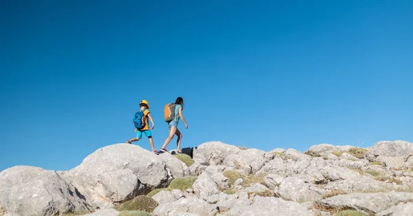 A woman and a child are walking along a mountain path. A boy with a backpack travels with his mother. Hiking with children. The kid is walking along the mountain range. Active summer vacation.