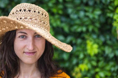 Hasır şapkalı gülümseyen bir kız sarmaşıkla kaplanmış bir duvarın yanında duruyor. Siyah saçlı bir kadının portresi.