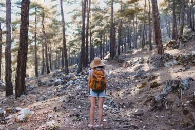 Ormanda sırt çantası ve hasır şapkalı bir kadının portresi, yürüyüş yapan gülümseyen bir kız, yaz yürüyüşü yapan, dağ yolunda yürümekten zevk alan mutlu bir kadın.