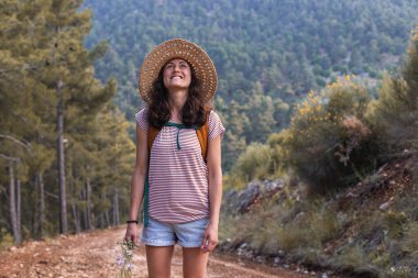 Ormanda sırt çantası ve hasır şapkalı bir kadının portresi, yürüyüş yapan gülümseyen bir kız, yaz yürüyüşü yapan, dağ yolunda yürümekten zevk alan mutlu bir kadın.
