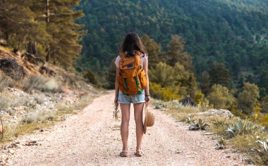 Sırt çantası ve hasır şapkalı bir kız büyük bir dağın arka planında, genç bir kadın dağ yolunda yürüyor, dünyanın en güzel yerlerine doğru bir yolculuk.