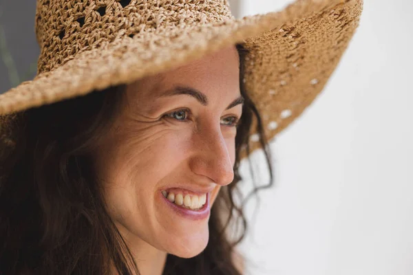 Geniş kenarlı hasır şapkalı güzel bir kadının portresi kameraya bakarken, koyu kıvırcık saçlı, gülümseyen ve yazın tadını çıkaran mutlu olgun bir kadının yüzünü kapat..