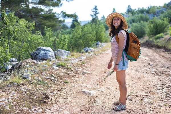 Ormanda sırt çantası ve hasır şapkalı bir kadının portresi, yürüyüş yapan gülümseyen bir kız, yaz yürüyüşü yapan, dağ yolunda yürümekten zevk alan mutlu bir kadın.