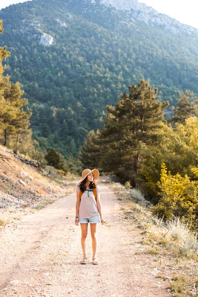 Sırt çantası ve hasır şapkalı bir kız dağ yolunda yürüyor, dağlarda yürüyor, bir kadın orman yolunda yürüyor, dünyanın en güzel yerlerine doğru bir yolculuk yapıyor.