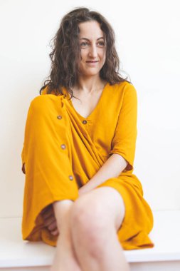 Sarı elbiseli gülümseyen kız beyaz bir duvarın arkasında oturuyor, kıvırcık saçlı bir kadının portresi.