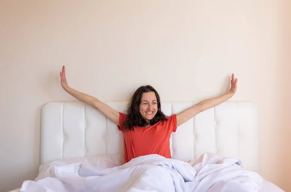 The girl sits on the bed and stretches, the woman woke up, a portrait of a brunette in a red T-shirt who sits on a white bed