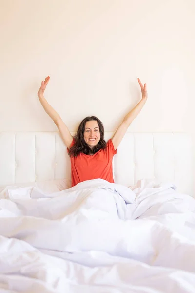 The girl sits on the bed and stretches, the woman woke up, a portrait of a brunette in a red T-shirt who sits on a white bed