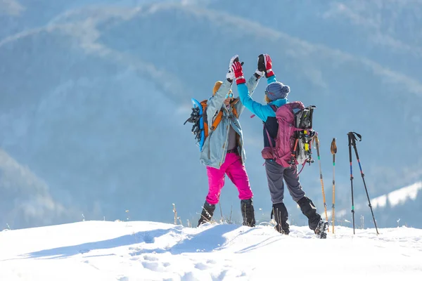 two women climbed to the top of the mountain during a winter hike, winter trekking, two girlfriends travel together, snow-capped mountains, girl gives high five to friend