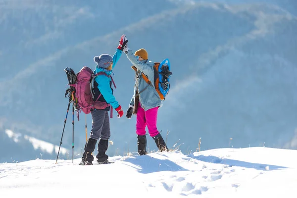 two women climbed to the top of the mountain during a winter hike, winter trekking, two girlfriends travel together, snow-capped mountains, girl gives high five to friend