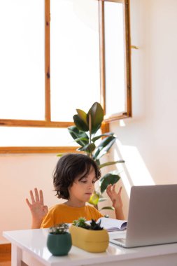 Uzaklık Eğitimi, okul çocuğunun portresi, öğretmenle video görüşmesi, çocuk masaya oturur ve parmaklar, jestler için ısınır, çocuk dizüstü bilgisayara bakar.