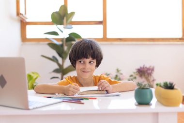 a boy writes and draws in a notebook during an online lesson. Distance online learning. a boy with a computer communicates via videoconference with a teacher and a class group. The child communicates online.
