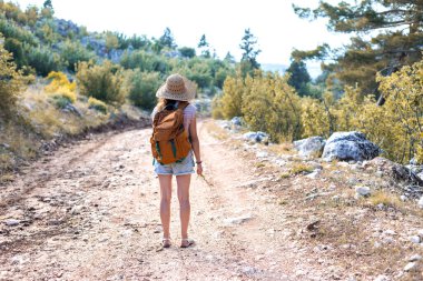 Ormanda sırt çantası ve hasır şapkalı bir kadının portresi, yürüyüş yapan gülümseyen bir kız, yaz yürüyüşü yapan, dağ yolunda yürümekten zevk alan mutlu bir kadın.