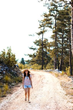 Ormanda sırt çantası ve hasır şapkalı bir kadının portresi, yürüyüş yapan gülümseyen bir kız, yaz yürüyüşü yapan, dağ yolunda yürümekten zevk alan mutlu bir kadın.