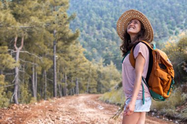 Ormanda sırt çantası ve hasır şapkalı bir kadının portresi, yürüyüş yapan gülümseyen bir kız, yaz yürüyüşü yapan, dağ yolunda yürümekten zevk alan mutlu bir kadın.
