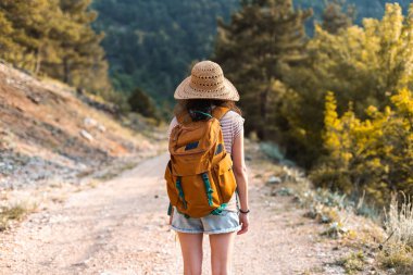 Sırt çantası ve hasır şapkalı bir kız dağ yolunda yürüyor, dağlarda yürüyor, bir kadın orman yolunda yürüyor, dünyanın en güzel yerlerine doğru bir yolculuk yapıyor.