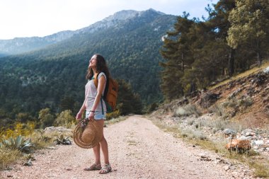 Dağ yolunda sırt çantalı bir kadın, dağlarda bir yürüyüş, orman yolunda yürüyen şapkalı bir kız, gülümseyen bir esmer