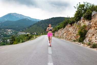 Bir kız asfalt bir yolda koşuyor güzel dağların arka planında, bir kadın akşam koşusunda, aktif bir yaşam tarzı, ince bir esmer dağ yolunda koşuyor.