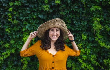 Hasır şapkalı gülümseyen bir kız sarmaşıkla kaplanmış bir duvarın yanında duruyor. Siyah saçlı bir kadının portresi.