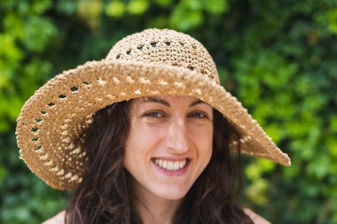 Geniş kenarlı hasır şapkalı güzel bir kadının portresi kameraya bakarken, koyu kıvırcık saçlı, gülümseyen ve yazın tadını çıkaran mutlu olgun bir kadının yüzünü kapat..