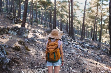 Ormanda sırt çantası ve hasır şapkalı bir kadının portresi, yürüyüş yapan gülümseyen bir kız, yaz yürüyüşü yapan, dağ yolunda yürümekten zevk alan mutlu bir kadın.