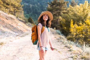 Dağlarda yürüyüşe çıkmış sırt çantalı bir kadın, orman yolunda yürüyen şapkalı bir kız, gülümseyen bir esmer