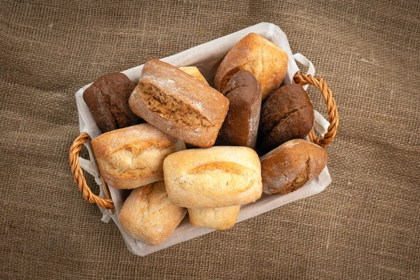 Tasty wheat and rye buns in a wicker basket on a burlap cloth — Stock Photo, Image