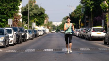 Happy middle aged woman jogging along a street with standing cars on the roadsides in the morning in Israeli city