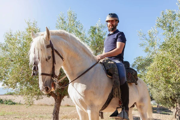 Kasklı bir binici, sakallı bir beyaz adam, arka planda zeytin ağaçları ve mavi gökyüzü ile çevrili bir alanda beyaz atına biniyor..
