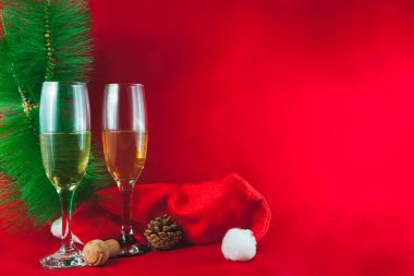 Kırmızı arka planda şampanya, çam kozalağı, tıpa, ağaç dalı ve Noel Baba şapkası olan iki kristal bardak var. Bir tarafta bir sürü kopya alanı var..