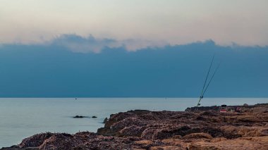 Deniz kıyısında, kayalık bir yüzeyde, bir masanın yanında iki olta, bir sandalye ve diğer balıkçı aletleri vardır. Arka planda, ufukta bulutlu bir gökyüzü görülebilir..