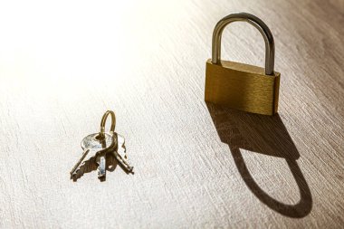 Ahşap bir masanın üzerinde bir asma kilit ve yanında üç anahtarlı bir yüzük var. Işık yukarıdan gelir ve masanın üstüne sert, uzun bir gölge bırakır..