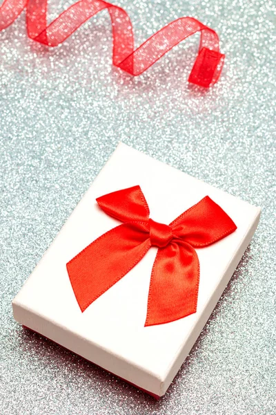 银光闪闪的表面是一个有白色盖子和红色蝴蝶结的礼品盒 旁边是一条波浪形的红丝带 没有对焦 这个图像是垂直方向的 — 图库照片